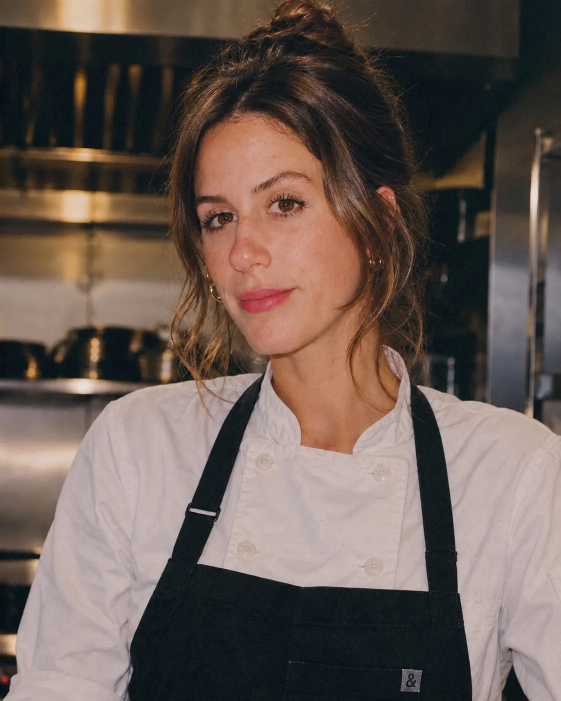 Chef Kristi in a white chef's coat and black apron, standing in the kitchen
