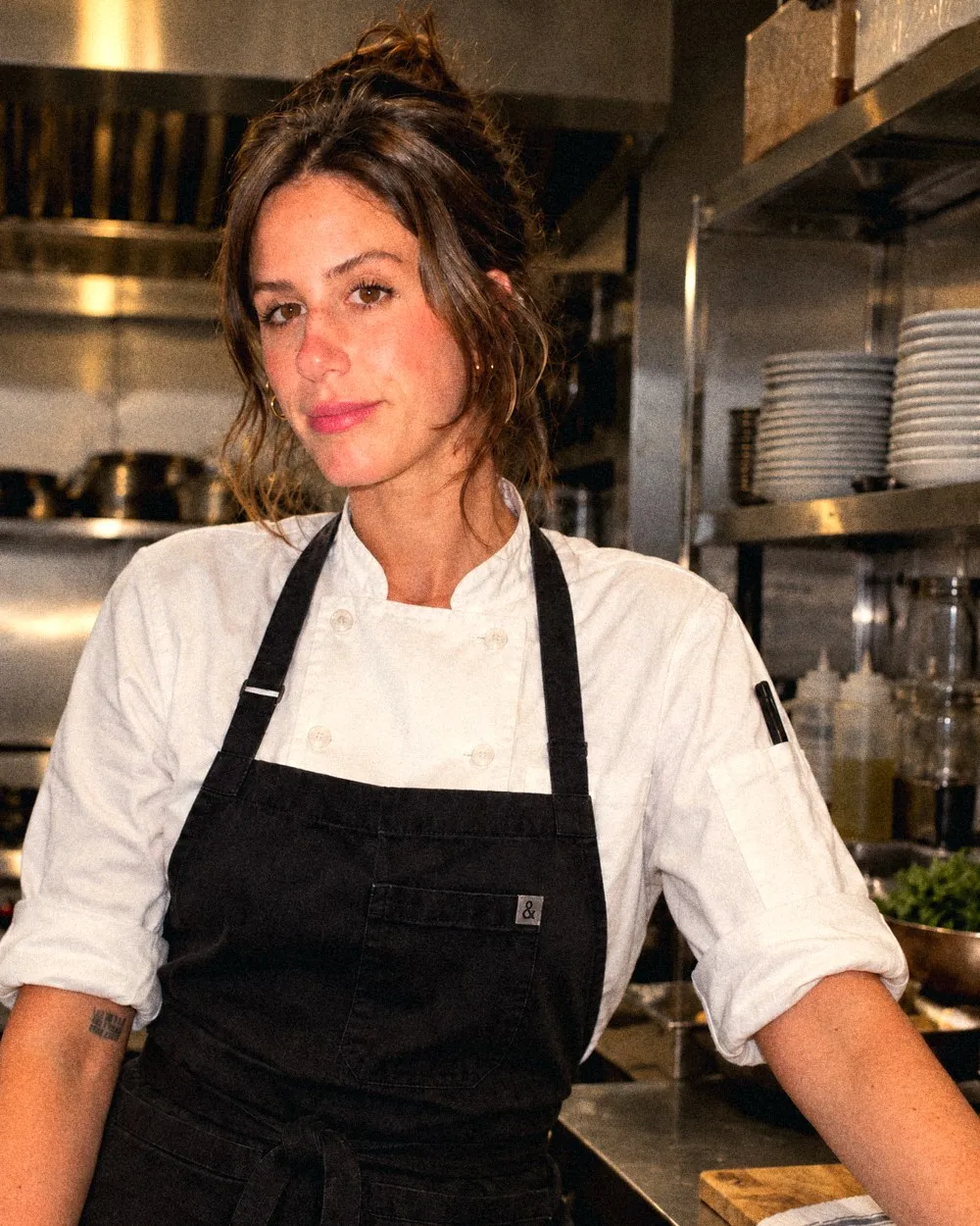 Chef Kristi in a white chef's coat and black apron, standing in the kitchen