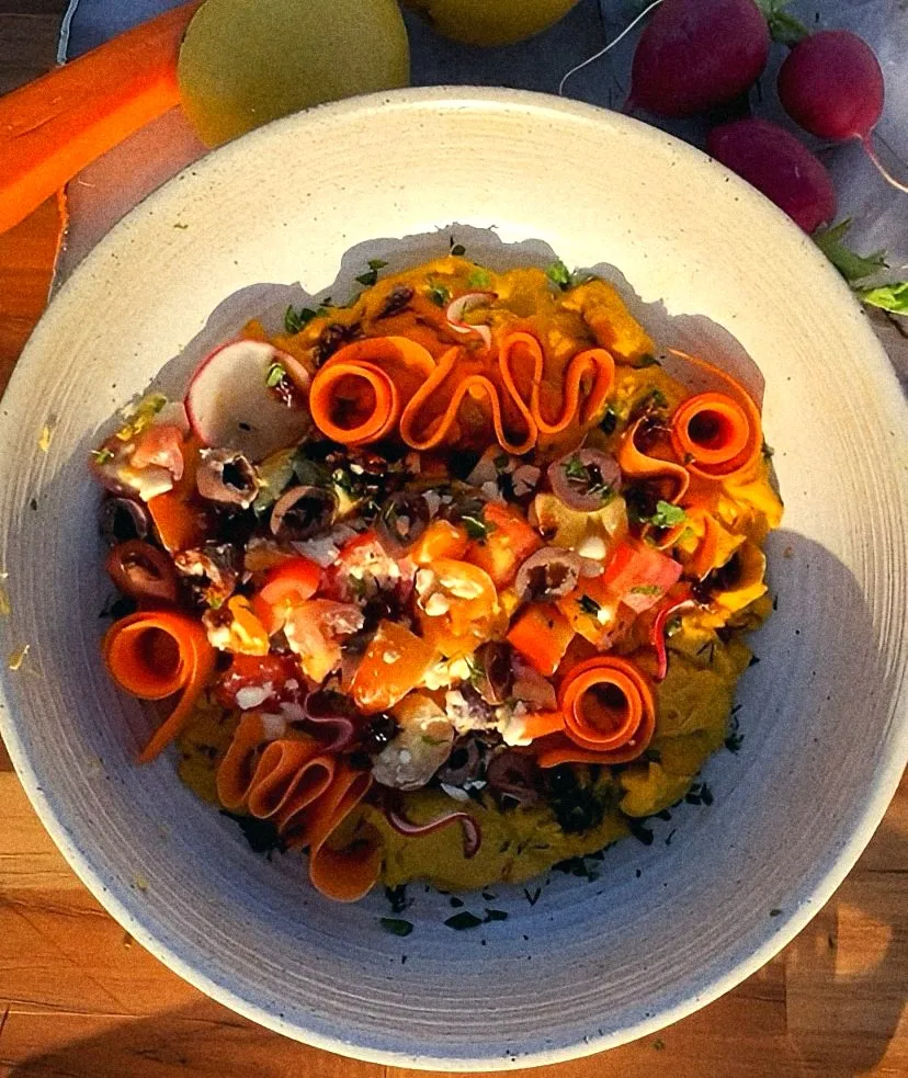 Turmeric hummus with carrot ribbons, olives, tomatoes and radish in a speckled bowl