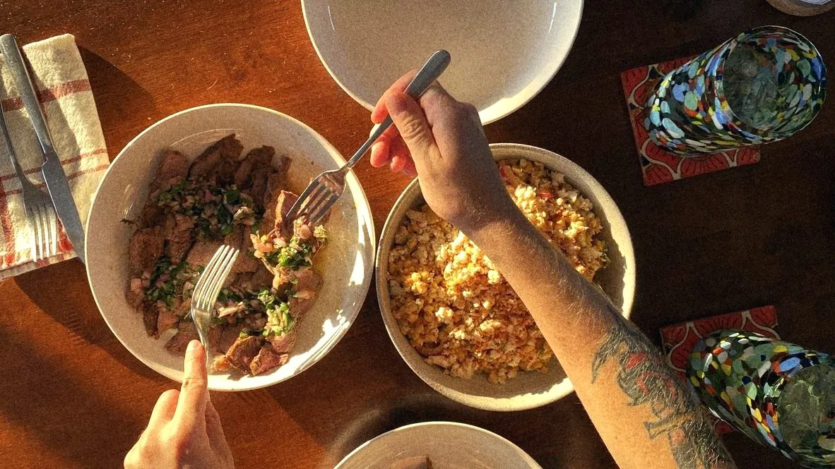 Hands reaching for sliced steak and street corn at a sunlit table