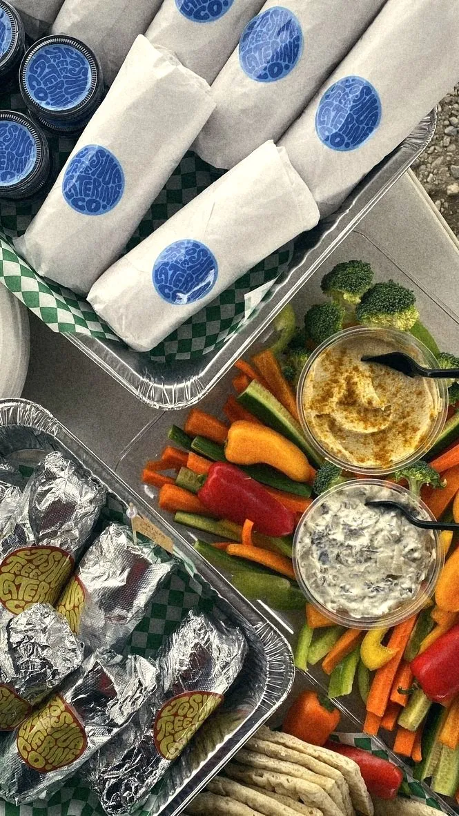 Wrapped sandwiches with JTCC stickers beside a crudité tray with hummus and dip